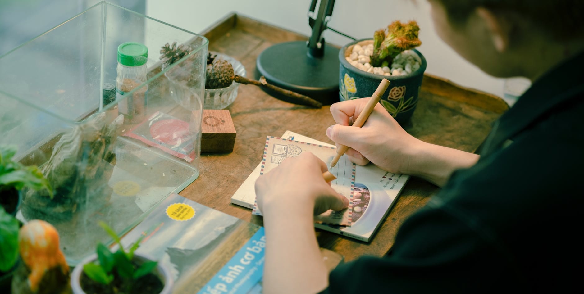 Osoba pisze wakacyjną pocztówkę przy drewnianym biurku z roślinami